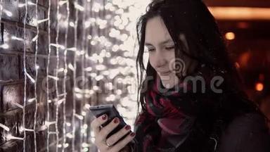 年轻迷人的女人在圣诞夜的大<strong>雪中</strong>使用智能手机<strong>站在</strong>灯光墙附近，
